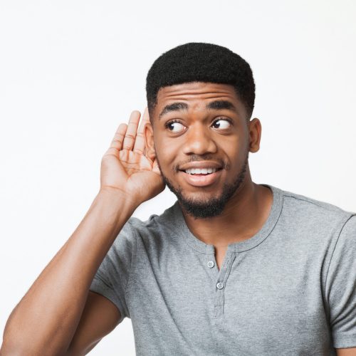 Curious interested african-american man holding hand near ear and trying to hear information, white studio background